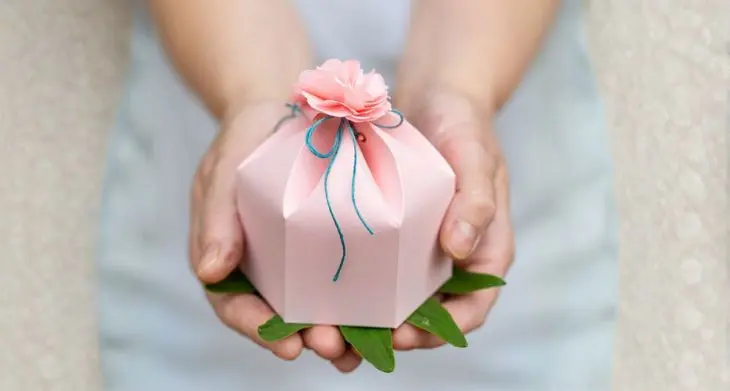 Detailaufnahme: Zwei Hände strecken eine selbstgebastelte Geschenkbox in rosa in die Kamera.