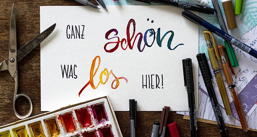 Im Handletteringstil steht „Ganz schön was los hier!“ auf einem Blatt Papier, drumherum liegen verschiedene Stifte.