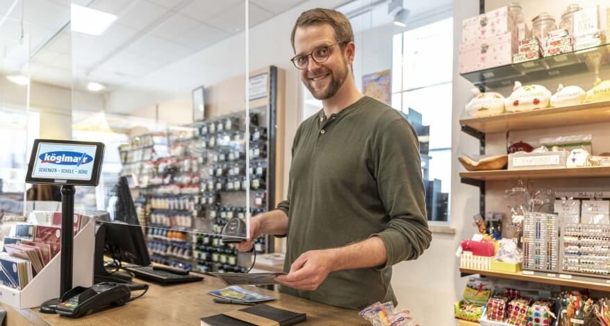 Ein Mann steht an einer Kasse und kassiert per Scanner einen Artikel. Im Hintergrund sind gefüllte Regale mit Bürobedarfsartikeln und Dekoartikeln zu sehen.