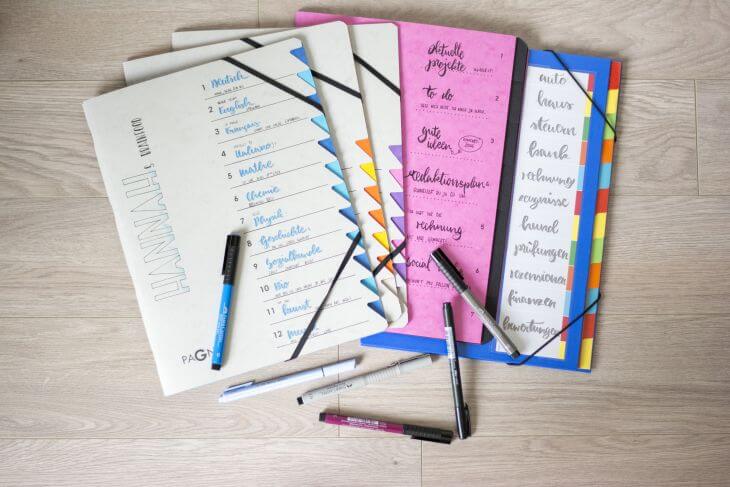 Auf einem Schreibtisch aus Holz liegen verschiedene Ordnungsmappen, die mit Handlettering beschriftet wurden. Darauf sind verschiedene Stifte verteilt.