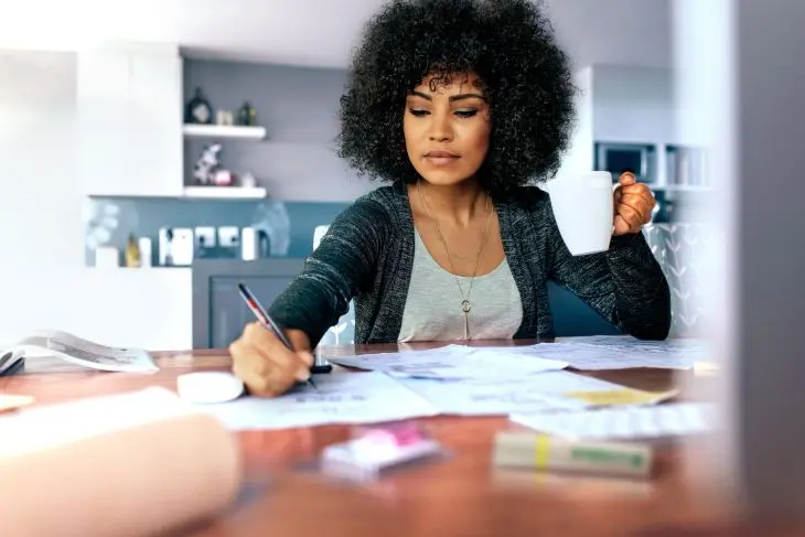 Eine Frau mit lockigem Haar sitzt an einem Tisch in einer modernen Küche. In der einen Hand hält sie eine weiße Tasse, in der anderen Hand schreibt sie mit einem Edding Fineliner auf vor ihr ausgebreiteten Blättern. 