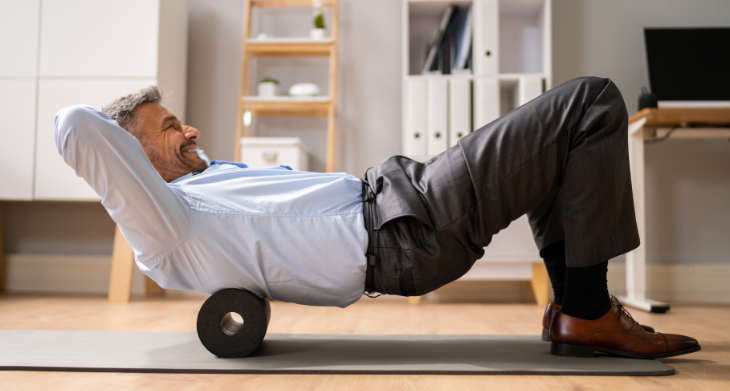 Das Bild zeigt einen Mann in Businesskleidung, der auf einer Yogamatte liegt und eine Blackroll zur Lockerung des Rückens verwendet. Er stützt seinen Oberkörper ab und rollt entspannt auf der Rolle, während er lächelt