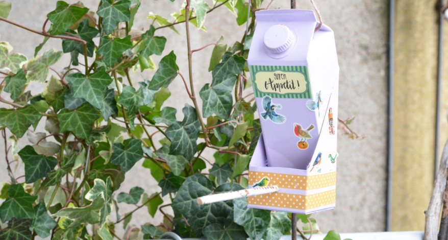 Ein selbstgebasteltes lila Vogelhäuschen, welches aus einem Milchkarton gebastelt wurde, hängt im Garten in einem Efeu-Strauch.