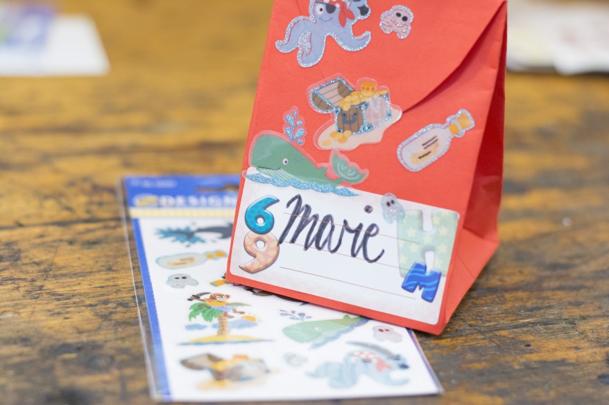 Eine rote Papiertüte in Form eines Schulranzens steht auf einem Holzboden. Diese wurde mit einigen maritimen Stickern von Avery Zweckform beklebt. Auf ein Etikett wurde der Name "Marie" gelettert. Unter der Tüte liegt eine gefüllte Packung Motivsticker von Avery Zweckform.