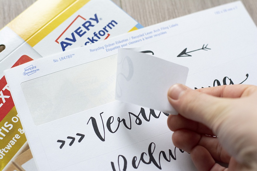 Eine Hand zieht ein bedrucktes Etikett für schmale Ordnerrücken von einem Bogen der Marke Avery Zweckform. Darunter liegt die Verpackung der Etiketten. 