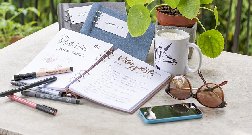Notizbücher, Stifte, Kaffee, Sonnenbrille und Smartphone draußen auf einem Tisch 