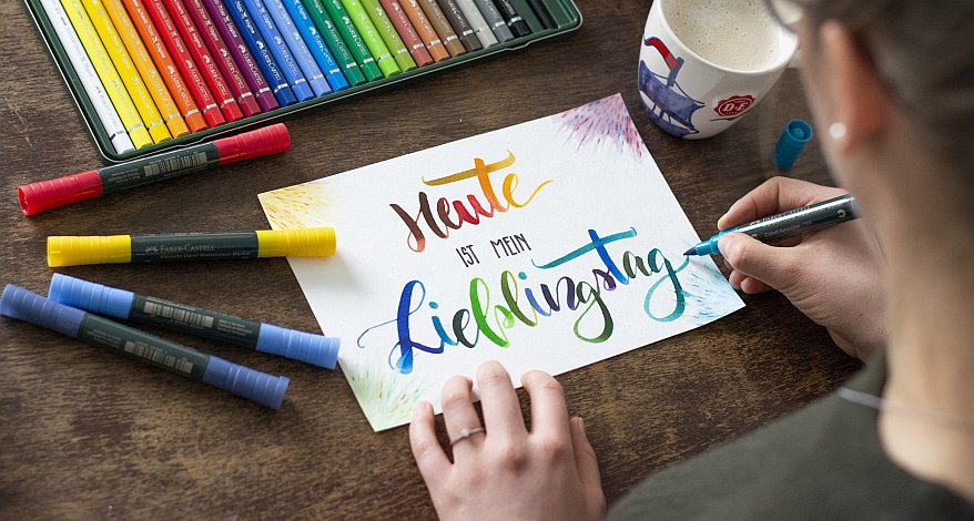 Jemand lettert in bunten Farben "Heute ist mein Lieblingstag" auf Papier, daneben bunte Stifte