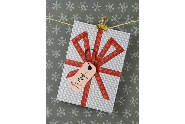 Eine grau-weiße Karte wurde mit rotem Washitape so beklebt, dass sie wie ein Geschenk aussieht. Darauf wurde ein Etikettensticker und Garn befestigt. Auf dem Etikett steht "Merry Christmas". Die Karte ist mit einer goldenen Foldbackklammer an einem Garn befestigt. Der Hintergrund ist grau und zeigt viele weiße Schneeflocken.
