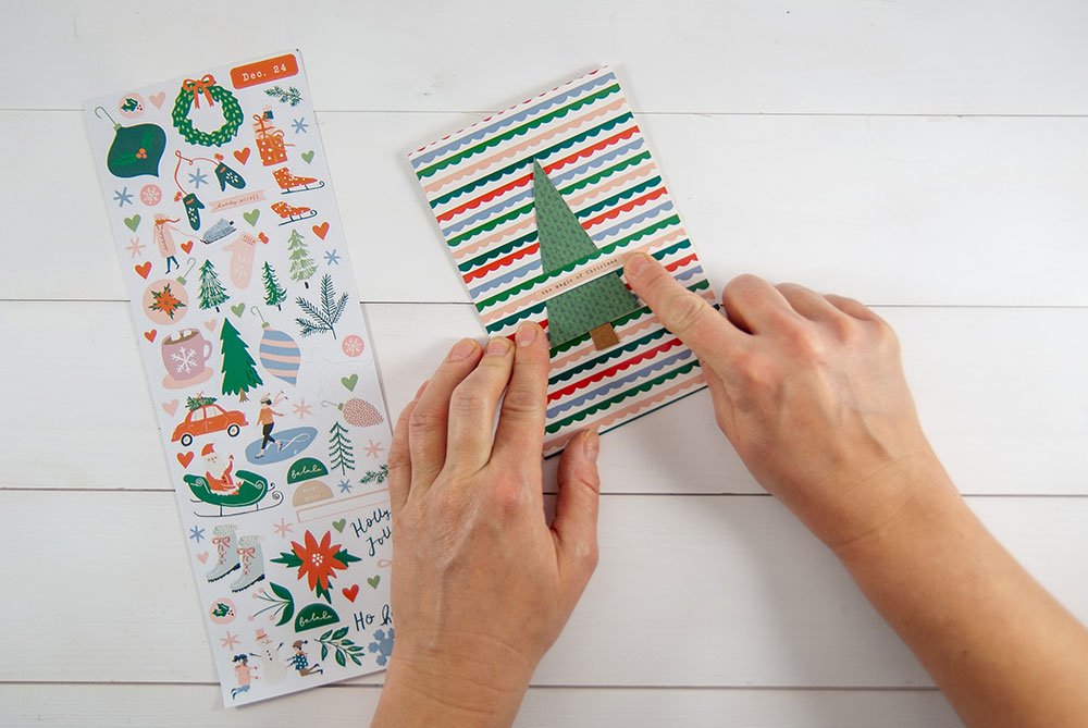 Eine Hand klebt einen Tannenbaum-Aufkleber der Marke Artoz auf eine Motivkarte, in die zuvor zwei Streifenschlitze geschnitten wurden. Am linken Bildrand liegt ein Stickerbogen mit zahlreichen Weihnachtsmotiven der Marke Artoz. 