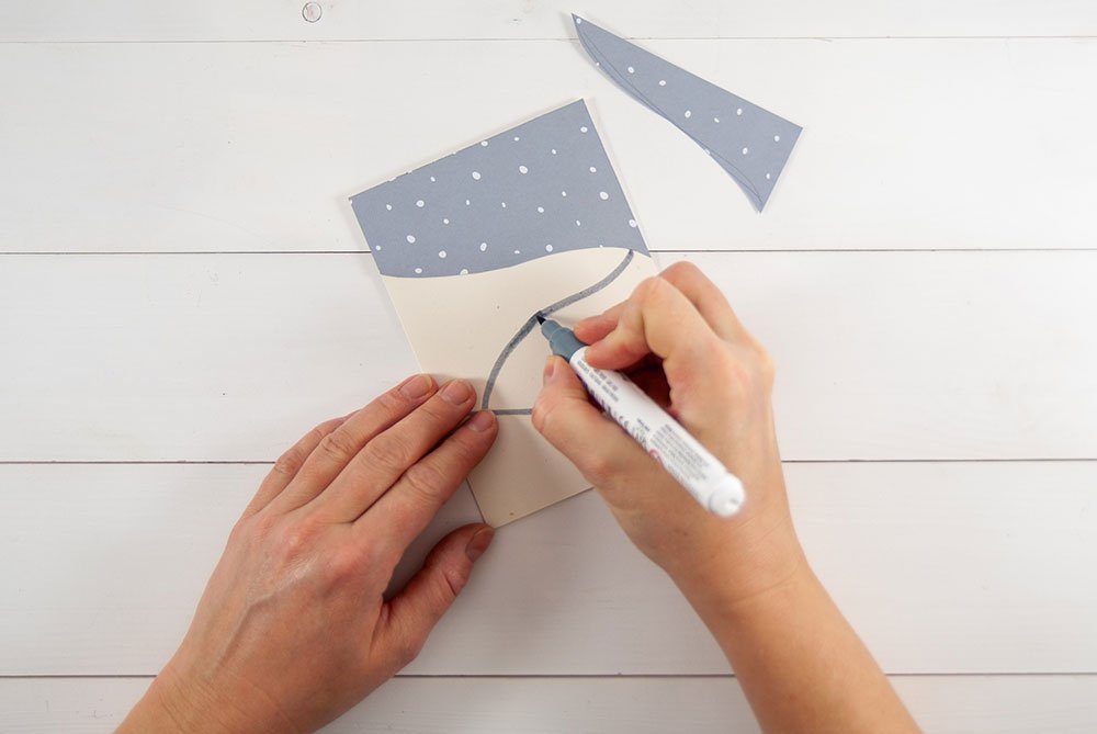 Auf einen weißen Karton wurde ein grauer Motivkarton geklebt, auf welchem mit weißen Marker Punkte als Schneeflocken gemalt wurde. Der graue Karton wurde dabei rund ausgeschnitten, so dass  der Eindruck eines Schneehügels entsteht. Eine Hand hält einen grauen Marker in der Hand, mit der graue Linien auf den weißen Karton gemalt werden.