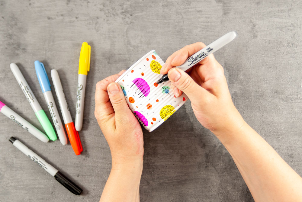 Eine Hand bemalt eine weiße Blechdose bunt, auf dem Tisch bunte Stifte.