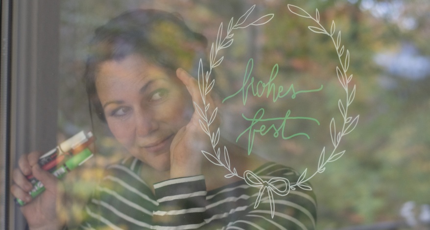 Eine junge Frau schaut durch ein Fernster, auf das sie mit Kreisemarkern desn Schridtzug "Frohes Fest" und einen Blumenkranz drumherum gemalt hat.