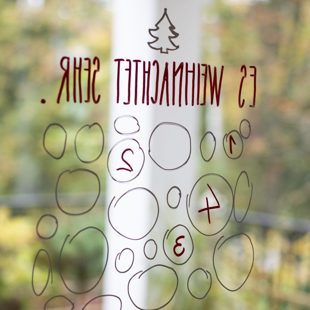 Mehrere Kreise wurden spiegelverkehrt auf ein Fenster gemalt und dienen akls Adventskalender. In drei Kreisen sind Zahlen geschrieben. Über den Kreisen steht "Es weihnachtet sehr" mit einem kleinen Tannenbaum.
