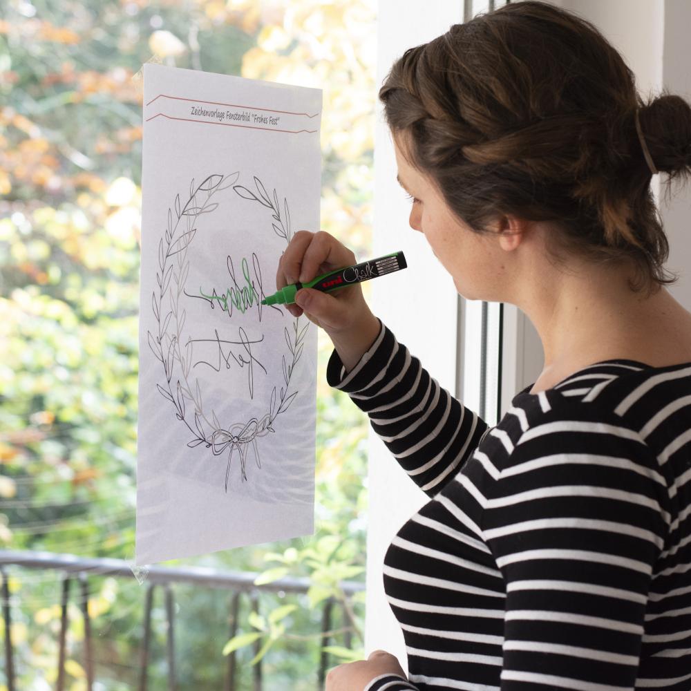Eine junge Frau hat die Malvorlage für das weihnachtliche Fensterbild von außen ans Fenster geklebt und malt nun mit einem grünen Kreidemarker von Uniball die Konturen nach.