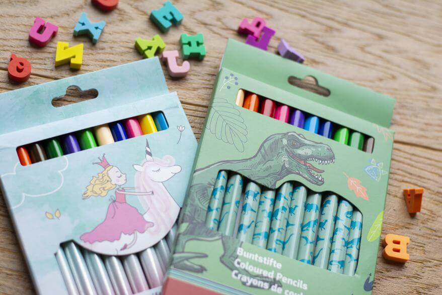 Zwei Buntstiftsets von Brunnen liegen auf einem Schreibtisch: Eines mit Einhornmotiv, eines mit Dinosauriermotiv. Dahinter liegen Radiergummis in ABC-Buchstabenform.