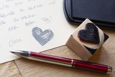 Nahaufnahme eines Briefes, dessen Ende mit einem gestempelten Herz versehen wurde. Daneben liegt ein roter Kugelschreiber "Pura" von Pelikan.