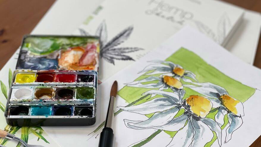 Ein Malblock aus Hanfpapier aus der Serie Natural Line von Hahnemühle liegt auf einem Tisch, daneben ein aufgekappter Farbkasten mit Aquarellfarben und eine Aquarellskizze mit Blumen. Foto und Illustration von Verena Knabe.