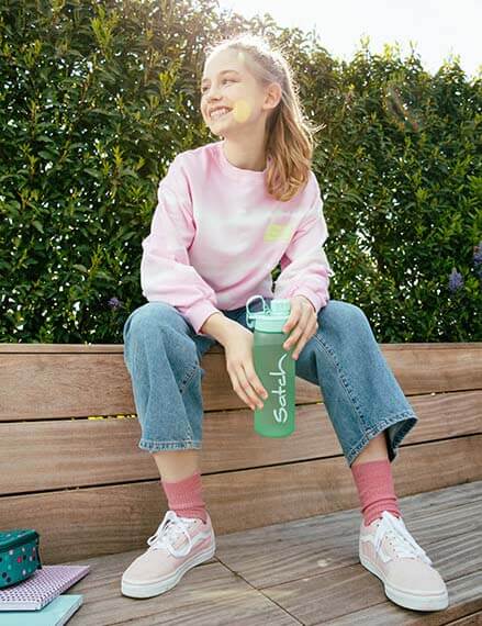 Ein Mädchen sitzt auf einer Bank und hält eine grüne Trinkflasche der Marke satch in den Händen. Vor ihr liegen einige Schulhefte und Schulbücher sowie ein Mäppchen.