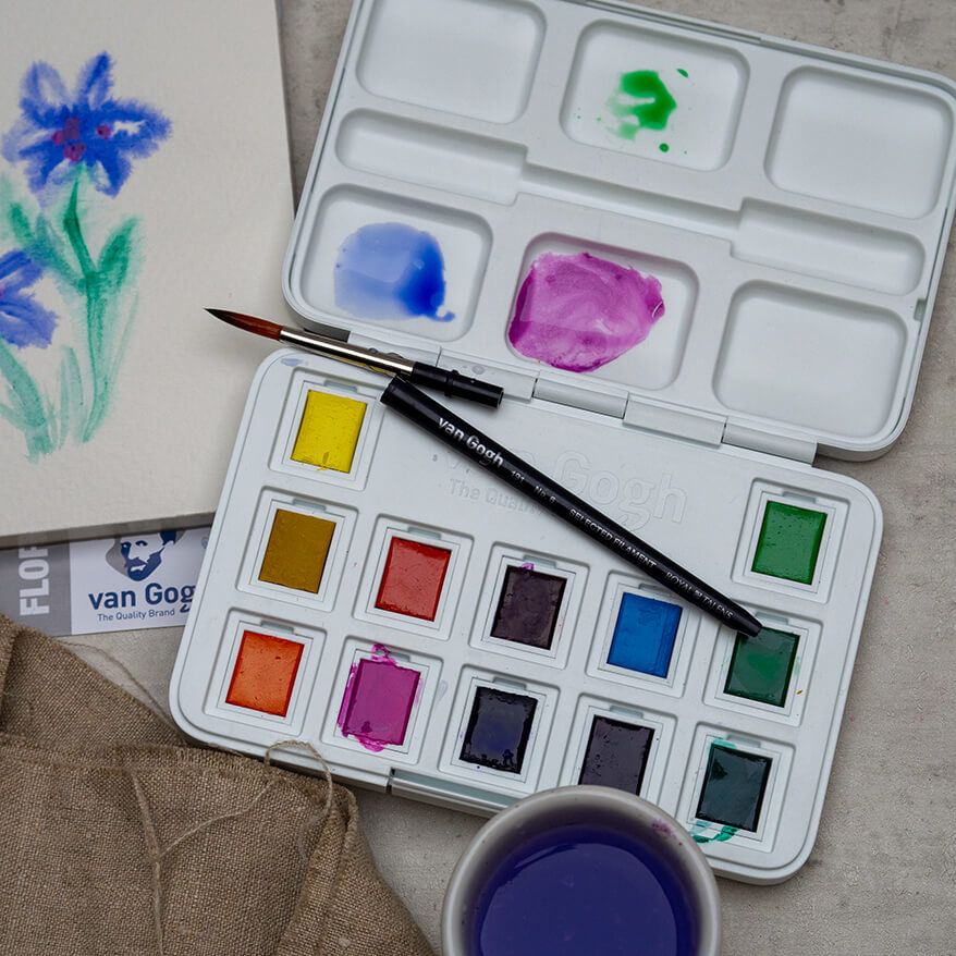 Auf einem grauen Untergrund liegt die Pocket Box “Frau Hölle” mit Aquarellfarben von Royal Talens mit einem zusammensteckbaren Mini-Pinsel. Außerdem ist ein Aquarell-Blumenbild, ein Wasserglas und ein Leinentuch zu sehen. Foto: Johanna Rundel