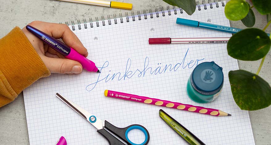 Eine Person schreibt mit der linken Hand mit Hilfe des Stabilo Tintenroller EASYoriginal das Wort "Linkshänder" auf einen Collegeblock. Auf dem Block liegen eine Linkshänderschere, ein Anspitzer für Linkshänder sowie weitere Stifte der Marke Stabilo. Im rechten Bildrand ist unscharf eine Pflanze zu erkennen. 