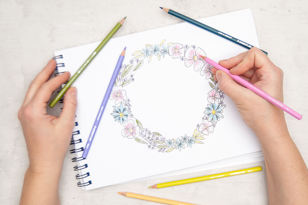 Jemand malt die Blüten und Blätter eines gezeichneten Blumenkranzes mit wasservermalbaren Buntstiften von Staedtler aus. Foto: Johanna Rundel