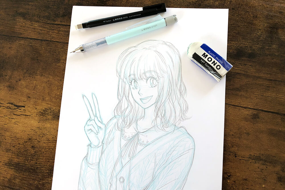 Auf einem braunen Holztisch liegt ein Blatt Papier. Darauf wurde mit Bleistift die Skizze einer jungen Frau im Manga-Stil gezeichnet. Außerdem sind folgende Materialien von Tombow auf dem Bild zu sehen: Radiergummi und Bleistifte. Foto: Reyhan Yildirim