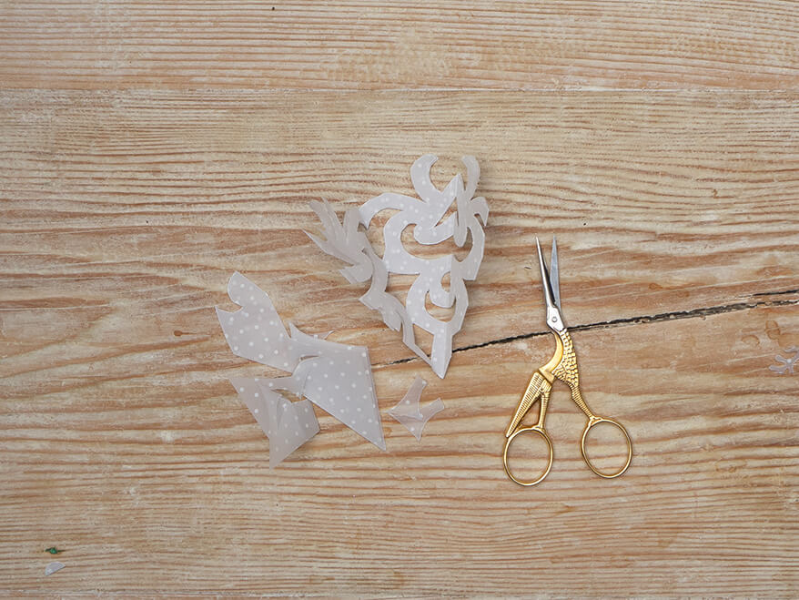 Aus Transparentpapier von Artoz wurde in Scherenschnitttechnik eine Schneeflocke ausgeschnitten. Die goldene Kranichschere sowie die Papierreste liegen daneben. Der Untergrund ist ein rustikales, helles Holzbrett. Foto: Johanna Rundel