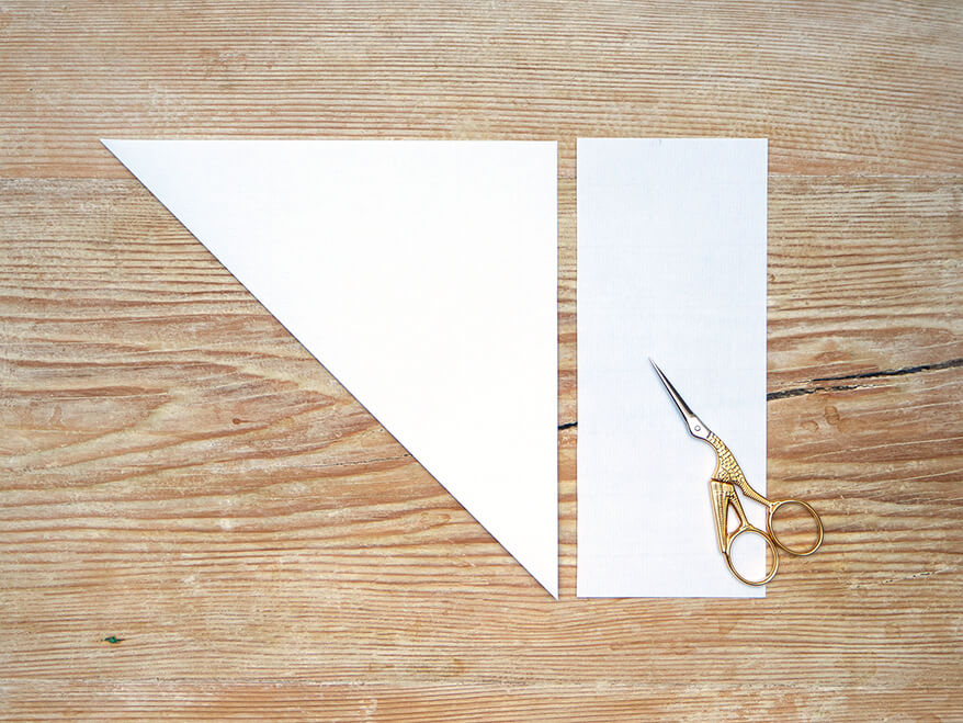Ein weißes Papier von Artoz im DIN-A4-Format wurde zum Dreieck gefaltet und der Überstand mit einer goldenen Kranichschere abgeschnitten. Der Untergrund ist ein rustikales, helles Holzbrett. Foto: Johanna Rundel