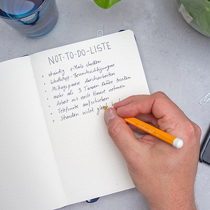 Jemand schreibt mit einem orangenen Drehgriffel eine Not-To-Do-Liste in ein Bullet Journal von Leuchtturm1917. Daneben ist ein Glas Wasser und eine Grünpflanze zu sehen. Einige weiße Büroklammern liegen verstreut darum. Der Untergrund ist hellgrau. Foto: Johanna Rundel 