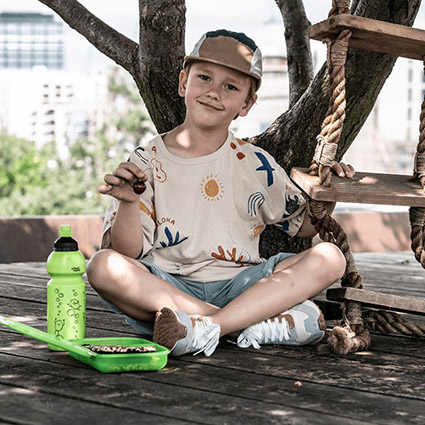 Ein Junge im Grundschulalter sitzt mit seiner grünen Trinkflasche und Brotdose von School-Mood in einem Baumhaus und hält sich mit der linken Hand an der Strickleiter fest. Mit der rechten Hand hält er eine Kirsche hoch. Der Junge ist sommerlich gekleidet und trägt eine Baseballmütze.