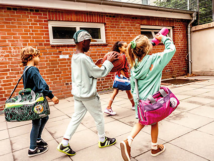 Vier Grundschulkinder laufen von links nach rechts an einem roten Backsteingebäude vorbei. Sie tragen alle Sporttaschen von School-Mood. Das blonde Mädchen vorne rechts trägt einen türkisfarbenen Pulli und trinkt aus einer pinken Trinkflasche. In der Mitte spielt ein Junge mit Baseballmütze mit einem Basketball.