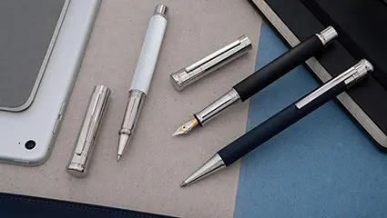 Verschiedene Schreibgeräte von Otto Hutt in schwarz und weiß liegen auf einem beige-blauen Untergrund. 