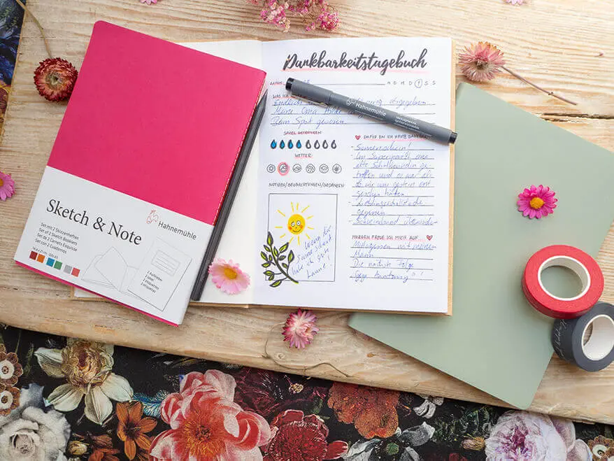 Auf einem rustikalen Holzbrett liegen verschiedene Skizzen- und Notizbücher von Hahnemühle. Eines ist aufgeschlagen. Die Seite eines Dankbarkeitstagebuchs ist zu sehen.