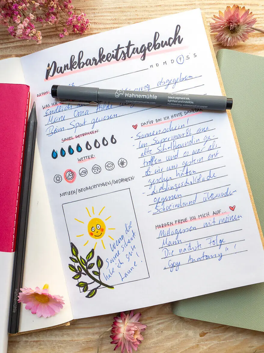 Auf einem rustikalen Holzbrett liegen verschiedene Skizzen- und Notizbücher von Hahnemühle. Eines ist aufgeschlagen. Die Seite eines Dankbarkeitstagebuchs ist zu sehen.