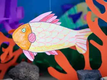 Ein großer, mit Buntstiften von STAEDTLER gestalteter Fisch aus Papier hängt in einem Aquarium aus einem Pappkarton. Foto: Johanna Rundel