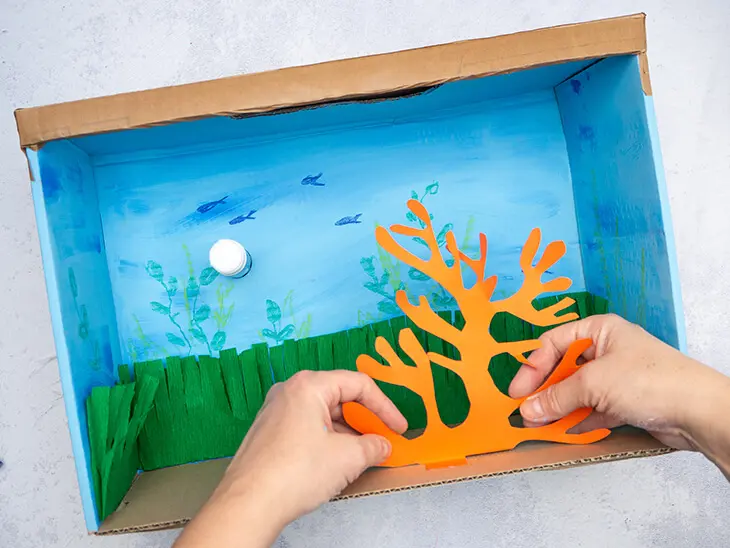 Jemand klebt eine Koralle aus Fotokarton in ein DIY-Aquarium aus Pappe. Foto: Johanna Rundel