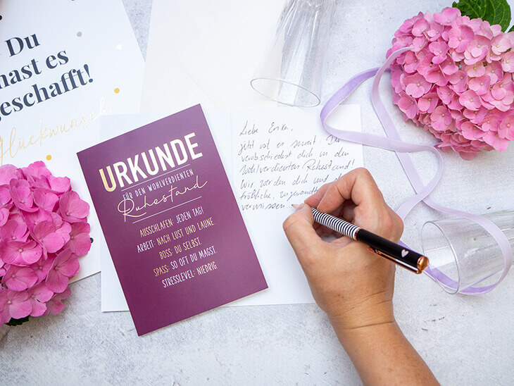Jemand schreibt einen Text in eine weinrote Karte zum Ruhestand von Grafik Werkstatt. Darauf steht abwechselnd in goldener und weißer Schrift: Urkunde für den wohlverdienten Ruhestand“. Foto: Johanna Rundel