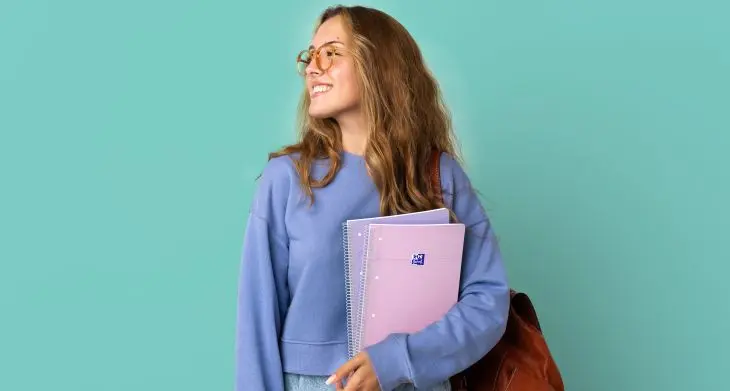 Vor einem türkisenen Hintergrund steht eine lächelnde Frau mit Collegeblöcken von Oxford im Arm