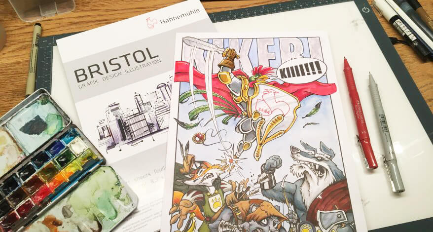 Blick auf einen Schreibtisch, auf dem ein koloriertes Comicbild liegt. Drumherum liegen Stifte, Bristolpapier von Hahnemühle und ein Aquarellkasten.