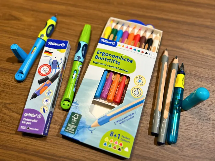 Mehrere Schreiblernstifte von Pelikan liegen auf einem Tisch, eine Kinderhand schreibt in ein Schulheft