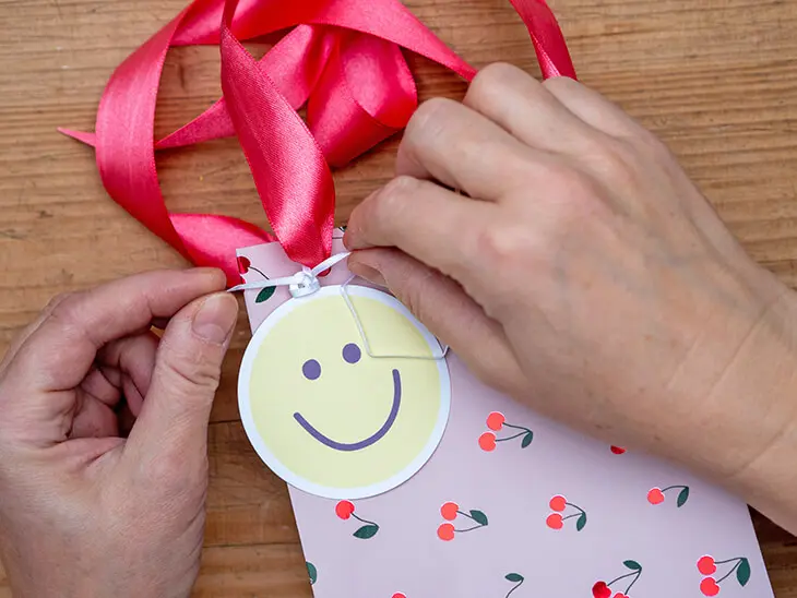 Jemand befestigt einen Smiley-Geschenkanhänger von STEWO an einer Geschenktüte. Foto: Johanna Rundel
