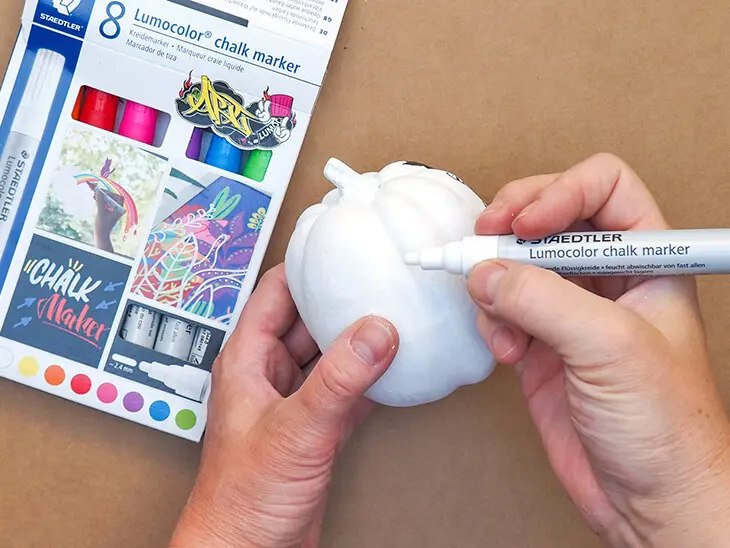 Jemand grundiert einen kleinen Kürbis mit einem weißen Chalk-Marker von STAEDTLER. Foto: Johanna Rundel
