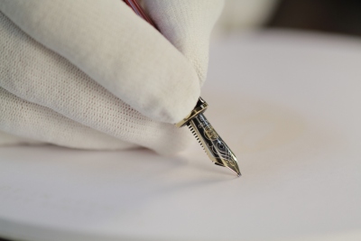 Eine Hand trägt einen weißen Samt-Handschuh. Diese Hand hält einen Füller der Marke Pelikan mit einer Goldfeder in der Hand und setzt ihn auf Papier auf.