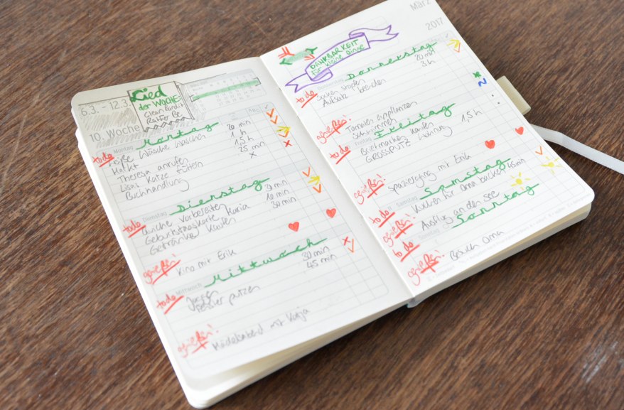 Auf einem Holztisch liegt ein aufgeschlagener Kalender mit vielen Notizen und Einträgen. Die Einträge sind bunt und kreativ gestaltet.