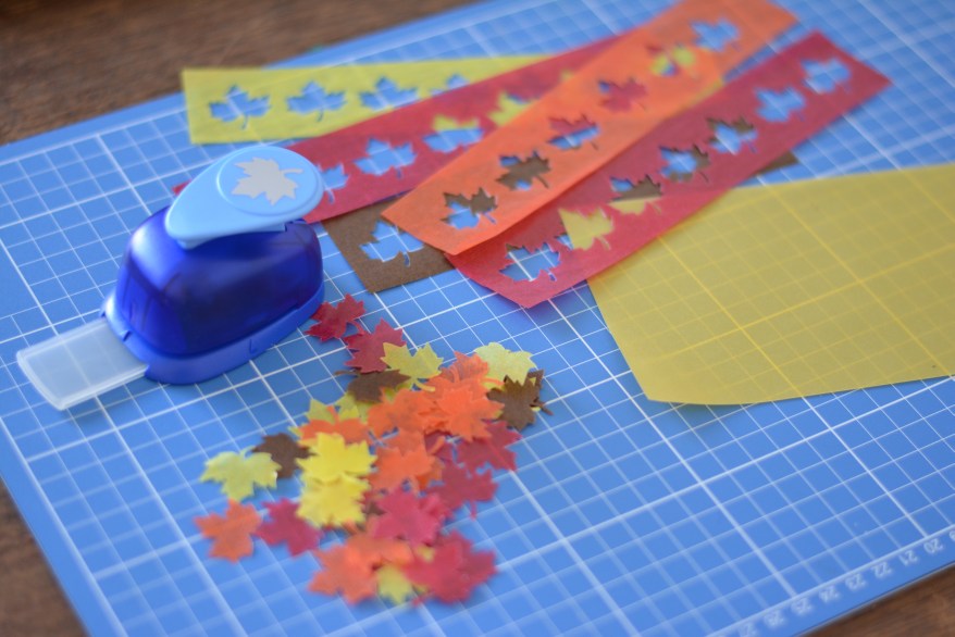 Auf einer Schneidematte liegen viele ausgestanzte Herbstblätter aus rotrem, gelben und orangem Transparentpapier. Daneben liegt der Stanzer der Marke Heyda.