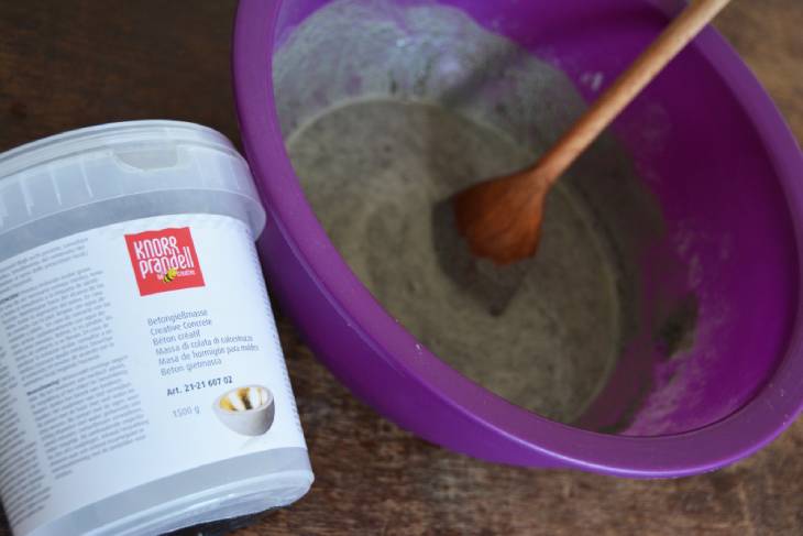 Auf einem Tisch steht eine lila Plastikschüssel, in der Bastelbeton angerührt ist. Daneben liegt ein Becher von Knorr Prandell mit Pulver für Bastelbeton.