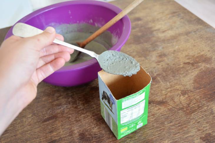 Auf einem Holztisch steht eine lila Plastikschüssel, gefüllt mit Bastelbeton. Mit einem Löffel wird der Bastelbeton in den die abgeschnittene Milchtüte gefüllt.