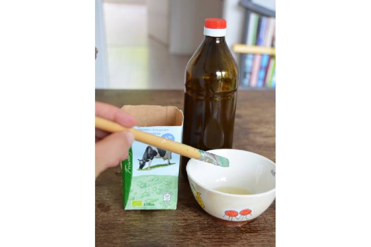 Auf einem dunklen Holztisch steht eine kleine Schüssel, in die Speiseöl gefüllt ist. Daneben steht eine Flasche Olivenöl und eine in der Mitte durchgeschnittene Milchtüte. Ein Pinsel tunkt in das Speiseöl.
