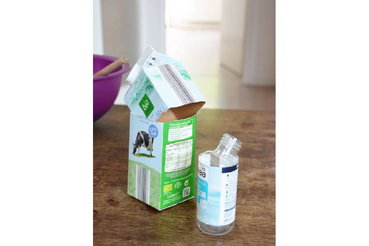 Auf einem Holztisch stehen eine aufgeschnittene Milchtüte und eine aufgeschnittene Plastikflasche.