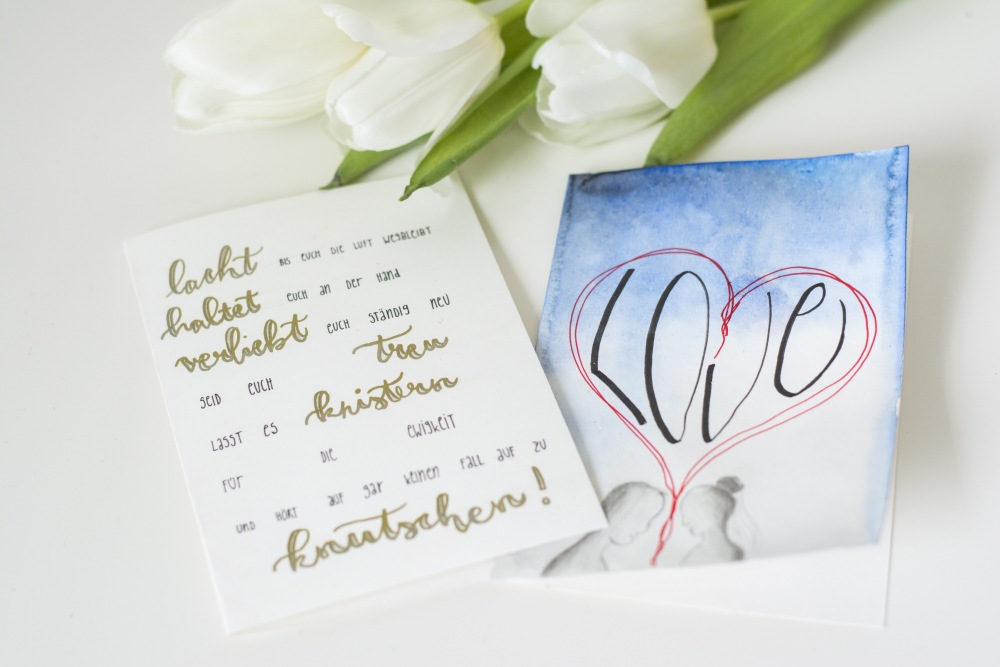 Auf einem weißen Tisch liegen 2 selbstgebastelte Hochzeitskarten. Die eine ist mit einem Spruch belettert, die andere ist mit einem Aquarellhintergrund in blau gestaltet. Darauf ist ein Herz und darin das Wort "love" gemalt, am unteren Rand ein Pärchen, das sich anschaut.
